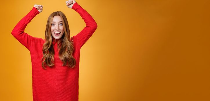 Optimistic happy and supportive ginger girl yelling cheering words, raising hands joyfully and smiling broadly triumphing, celebrating success and win, posing satisfied and excited against orange wall.