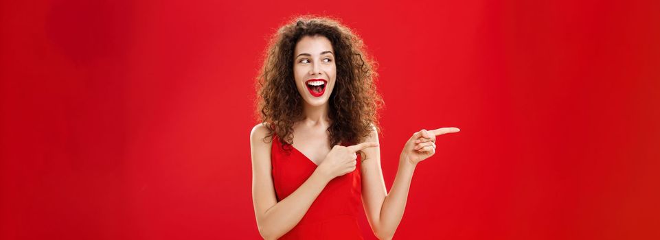 Waist-up shot of amused and entertained charming happy female with curly hairstyle in elegant evening dress and make-up pointing, looking left delighted and happy laughing joyfully over red background.