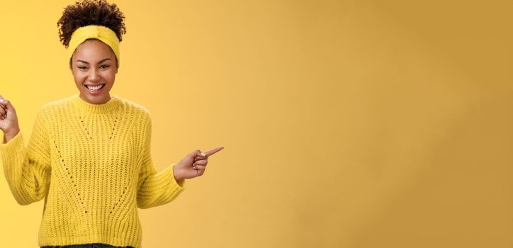 Charming tender romantic young african-american 20s woman smiling friendly satisfied laughing lovely look camera pointing up right sideways show products choices two ways, standing yellow background.