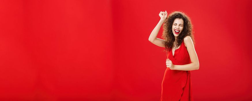 Rich woman forgetting all troubled on dance floor. Charming elegant young european female model in stylish evening dress moving to rhythm of music rejoicing raising hand, singing posing over red wall. Copy space