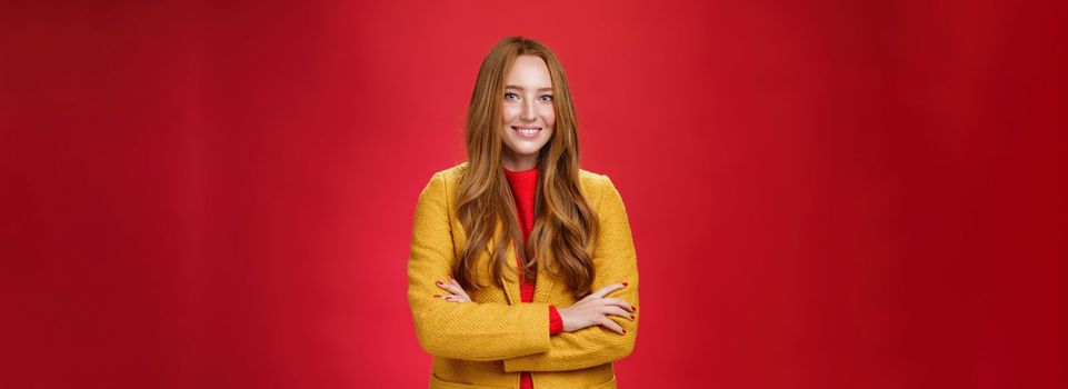 Lifestyle. Charming successful young female in yellow autumn coat smiling broadly holding hands crossed on body in confident self-assured pose standing delighted and happy over red background.