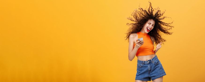 Portrait of joiyful carefree and happy charming european woman with curly hairstyle laughing out loud holding smartphone wearing wireless earbuds listening awesome music via app over orange wall. Technology concept