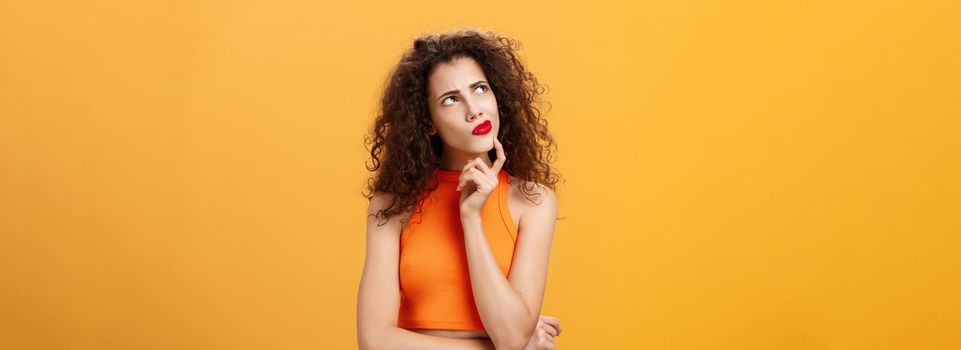 Waist-up shot of perplexed thoughtful. and smart female with curly hairstyle in red lipstick and cropped top holding in hmm pose touching chin frowning looking at upper left corner thinking determined.