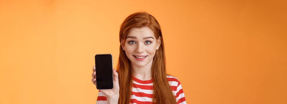 Close-up lovely caucasian redhead girl showing smartphone screen orange background, smiling cute, recommend application, promote app, online offer sale, introduce mobile phone. Technology concept