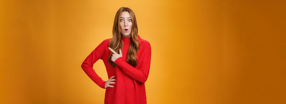 Impressed and surprised european ginger girl in red warm sweater open mouth from excitement and amazement pointing at upper right corner, gasping astonished and curious, asking question.