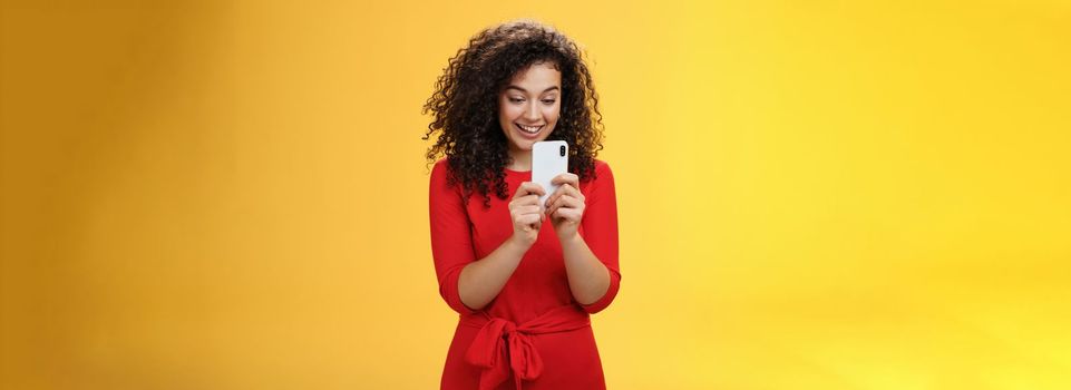 Wow new mobile phone amazing. Impressed and astonished good-looking curly-haired female in red dress holding smartphone looking at screen amused as playing cool app or game over yellow wall.