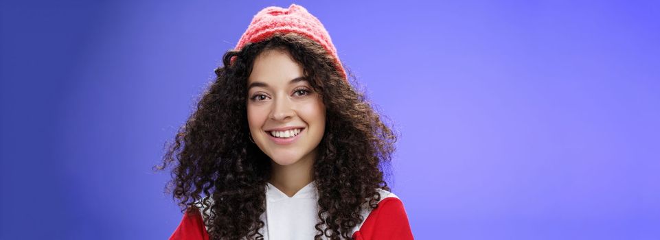 Headshot of tender and cute curly-haired 20s woman in warm beanie and cool sweatshirt smiling broadly enjoying awesome sunny and chilly days outdoors having fun posing over blue background. Copy space