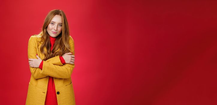 Romantic good-looking tender and cute ginger girlfriend. with freckles hugging herself and looking with sensual gentle smile at camera getting cold or chilly waiting outside over red background.