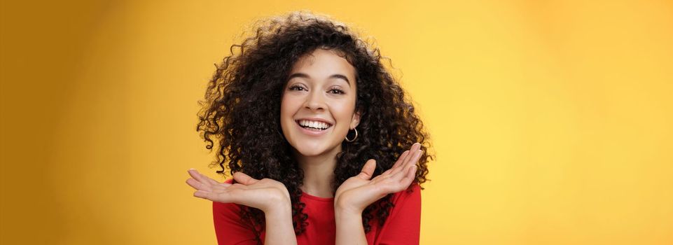 Lifestyle. Close-up shot of happy kind and tender pretty caucasian female student with curly hair and perfect skin smiling delighted holding palms spread near face having fun over yellow background.