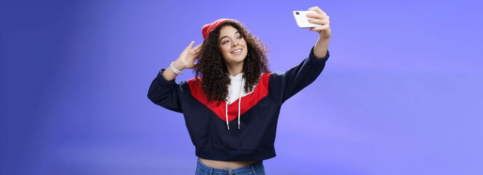 Cute girl making selfie to memorize trip standing in warm winter beanie and sweatshirt posing as pulling hand with smartphone and smiling at device screen to take pretty shot and post internet. Technology and lifestyle concept