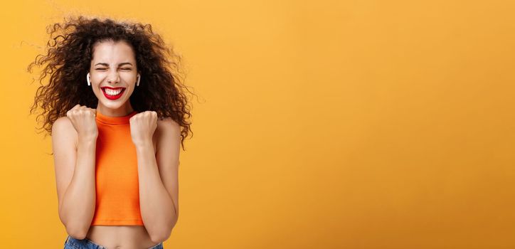 Pleased happy and triumphing curly-haired female in cropped top clenching fists in yes gesture closing eyes and smiling broadly receiving awesome news, winning award over orange background. Copy space