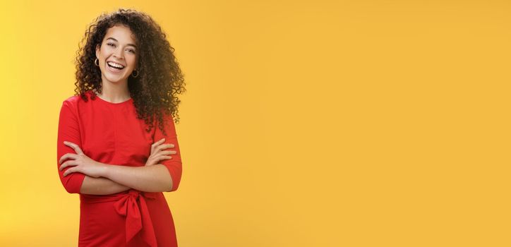 Self-assured happy enthusiastic curly-haired female reporter in cute red dress laughing carefree, having fun tilting head amused and holding hands crossed over body in confident pose over yellow wall.