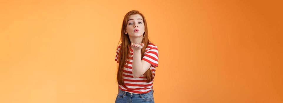 Silly cute and flirty redhead passionate girl, send air kiss camera, fold lips hold arm near mouth to blow mwah, romantic tender attitude, confess love like guy, stand orange background.