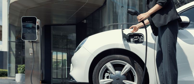 Closeup progressive suit-clad businesswoman with her electric vehicle recharge her car on public charging station in modern city with power cable plug and renewable energy-powered electric vehicle.