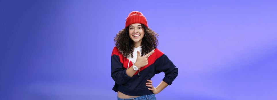 Studio shot of happy charming stylish city girl in beanie and warm sweatshirt ready rock winter up pointing at upper left corner and smiling delighted showing cool place over blue wall. Copy space