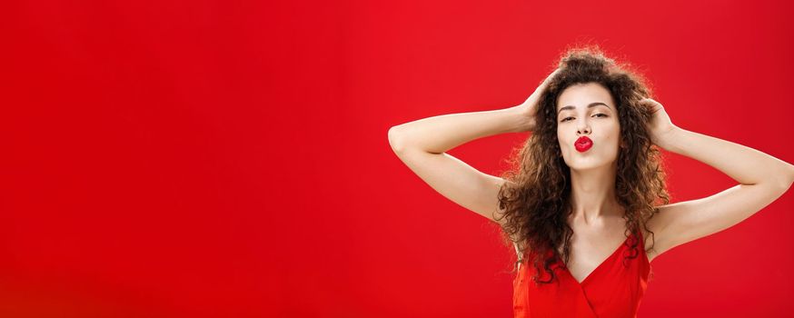 Waist-up shot of confident flirty and elegant young rich woman. with curly hairstyle in red lipstic and evening dress touching hair folding lips in mwah giving kiss to admirer feeling self-assured.