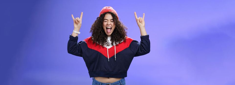 Waist-up shot of rebellious and carefree woman having fun feeling excited and awesome at party close eyes sticking out tongue and showing rock-n-roll signs, wearing beanie and warm clothes.