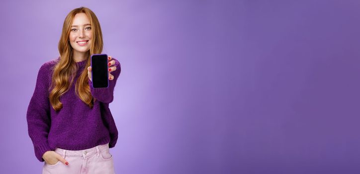 Lifestyle. Girl shows smartphone screen at camera to ask opinion of friend smiling broadly with optimistic and joyful expression holding hand in pocket promoting mobile phone or app over purple background.