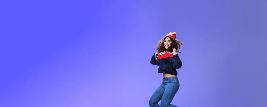 Studio shot of cute girl with curly hair in beanie sweatshirt and sneakers jumping playful and carefree over blue background, having fun enjoying cool weather smiling broadly as flying in air.