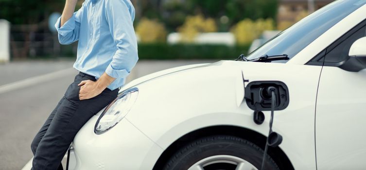 Closeup progressive suit-clad businessman with his electric vehicle recharge his car on public charging station in modern city with power cable plug and renewable energy-powered electric vehicle.