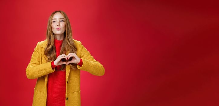 Love you all. Portrait of romantic and stylish good-looking flirty redhead female with freckles and blue eyes folding lips to give kiss showing heart gesture, confessing in sympathy over red wall.