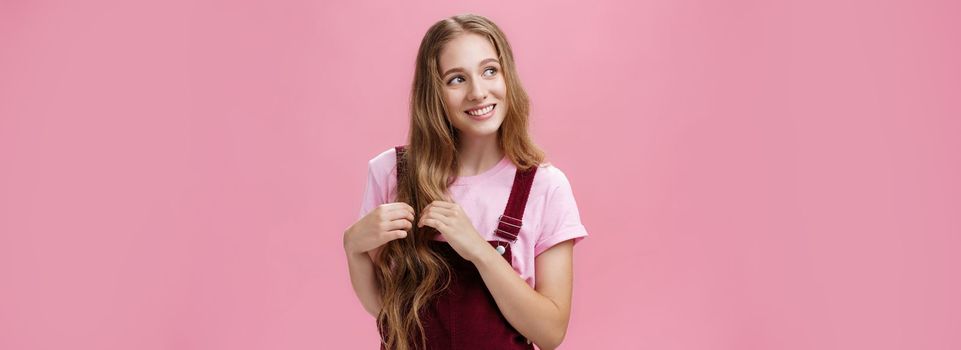 Silly kind and friendly-looking charming girl daydreaming picking strands of hair and looking left with nice cute smile imaging, picturing lovely scene posing flirty and happy over pink background.