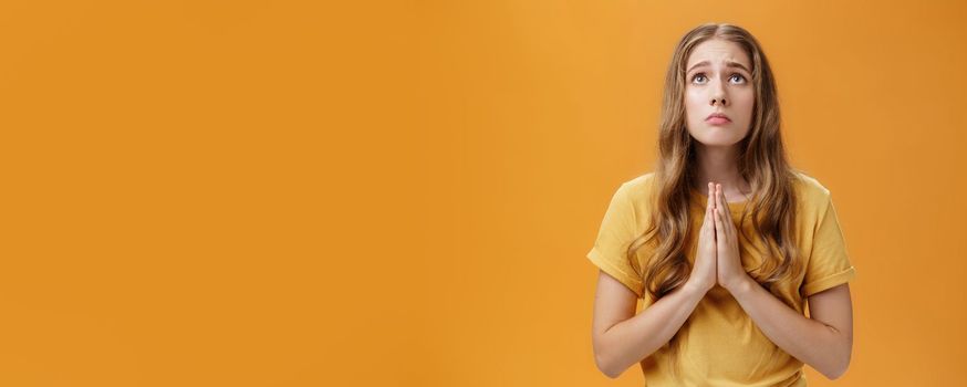Hopeful uneasy and worried gloomy faithful girl in t-shirt holding hands in pray against chest looking up with sad look praying making wish for good well of family posing over orange wall. Body language concept
