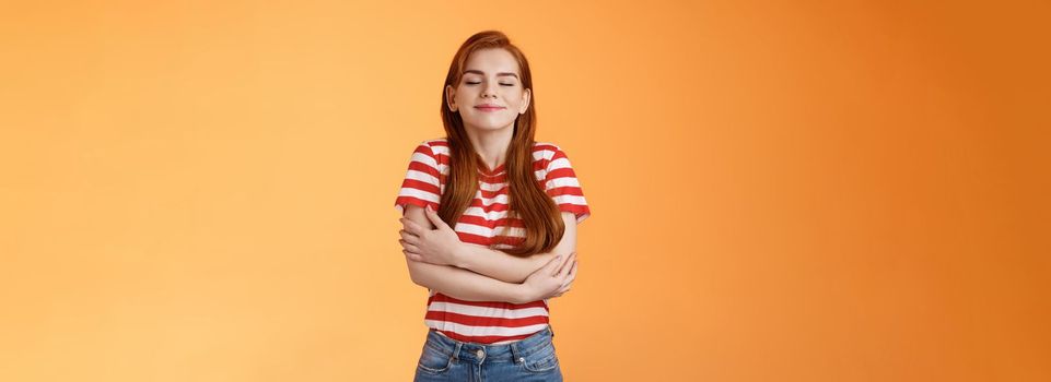 Tender lovely redhead modern woman embrace own body, feel happiness self-acceptance, smiling close eyes, cross arms, hugging herself dreamy, imaging something, daydreaming orange background.