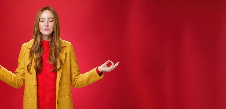 Girl keep calm releasing stress with meditation, posing in yellow coat, close eyes and looking relieved as extending hands sideways with mudra gesture, doing yoga against red background in lotus pose.