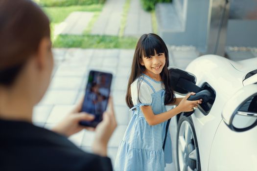 Progressive mother using smartphone take focus photo of daughter recharge EV car. Image of asian girl insert electric charger from home charging station to EV car on phone screen with blur background.
