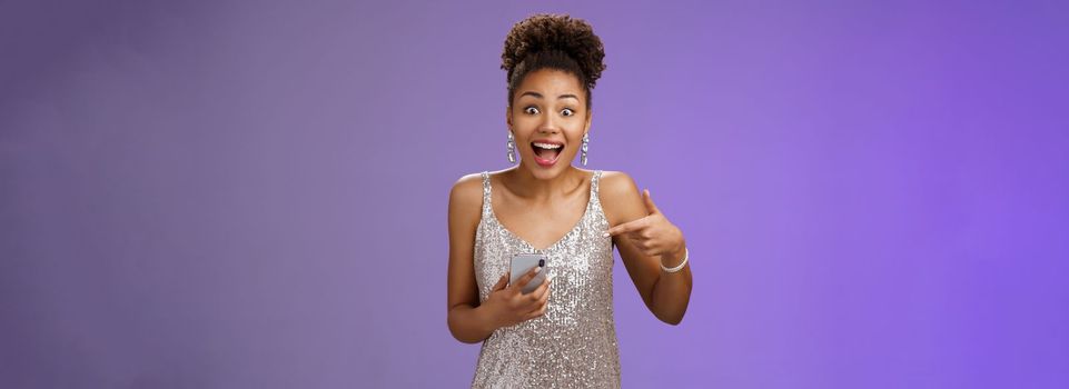 Amused charming glamour african-american woman in glittering silver dress astonished thrilled pointing smartphone receive incredible good perfect news telling you, standing blue background.