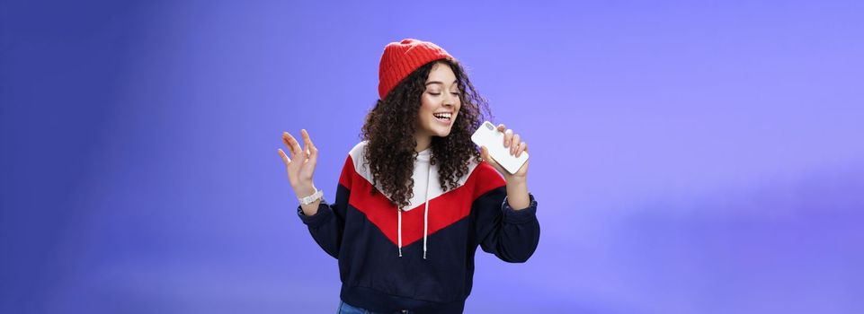 Attractive happy young woman with curly hair in hat singing enjoying perfect winter day singing along in smartphone, holding mobile phone like microphone, adore karaoke over blue background.