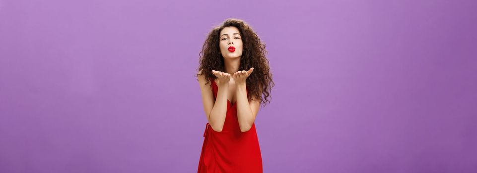 Tender charming european woman. with red lipstick in elegant evening dress bending towards camera folding lips sending air kiss at camera holding palms near mouth expressing love and passion.