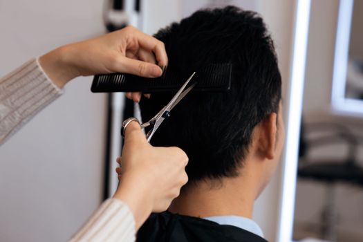 Closeup asian female barber making haircut for male customer with male hairstyle in qualified barbershop. Men's hairstyling by a scissor and comb in hair salon concept.