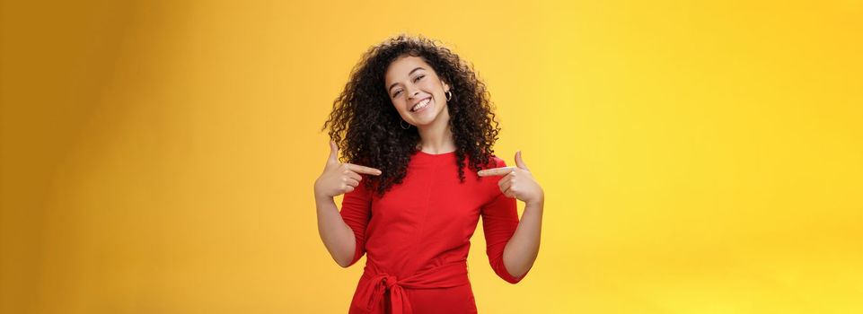 Lifestyle. Proud and satisfied ambitious successful female student in red dress standing pleased smiling and pointing at herself as if bragging about own achievements happily and glad over yellow wall.