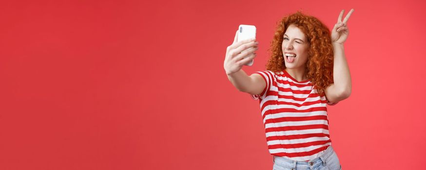 Lifestyle. Sassy fashionable playful good-looking redhead daring curly woman show peace victory animal ears gesture winking smartphone display record vlog taking selfie awesome phone camera red background.