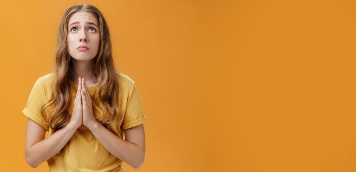 Hopeful uneasy and worried gloomy faithful girl in t-shirt holding hands in pray against chest looking up with sad look praying making wish for good well of family posing over orange wall. Body language concept