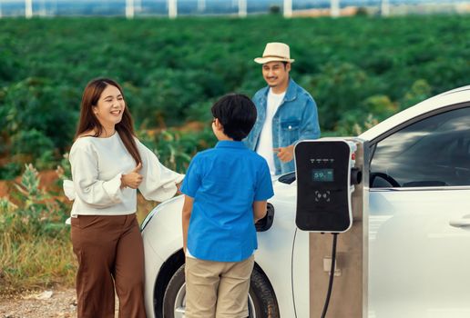Concept of progressive happy family enjoying their time at wind farm with electric vehicle. Electric vehicle driven by clean renewable energy from wind turbine generator for charging station.