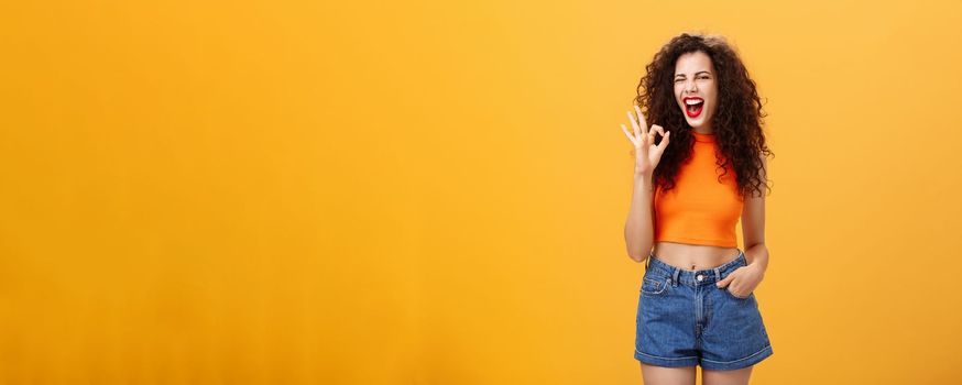 Excellent party like it. Joyful enthusiastic young curly-haired female with red lipstick in stylish cropped top winking excited smiling and showing okay or perfect sign posing over orange background.