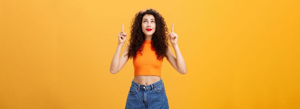 Woman wishing on shooting start believing in miracle looking and pointing hopefully up dreaming and praying in plan fullfillment standing in stylish urban cropped top over orange background. Copy space