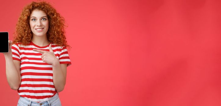 How about this app. Redhead curly friendly cute girl help friend find oufit online store hold smartphone pointing phone screen smiling broadly recommend social media photo filter, red background.