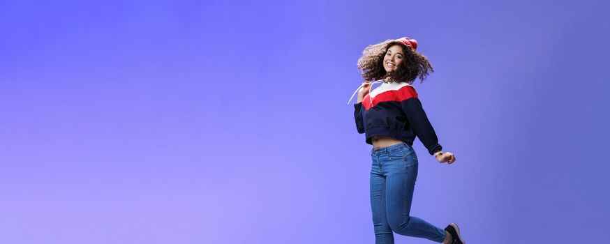 Dreamy and femenine cute girl with curly hair in winter beanie, jeans and sweatshirt jumping over blue background with satisfied carefree smile looking at camera feeling light and joyful.