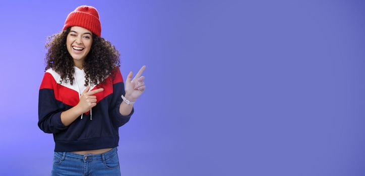 Charismatic playful young curly-haired european female winking joyfully at camera as pointing at upper right corner wearing hat and pullover as posing delighted over blue background, feeling warm.
