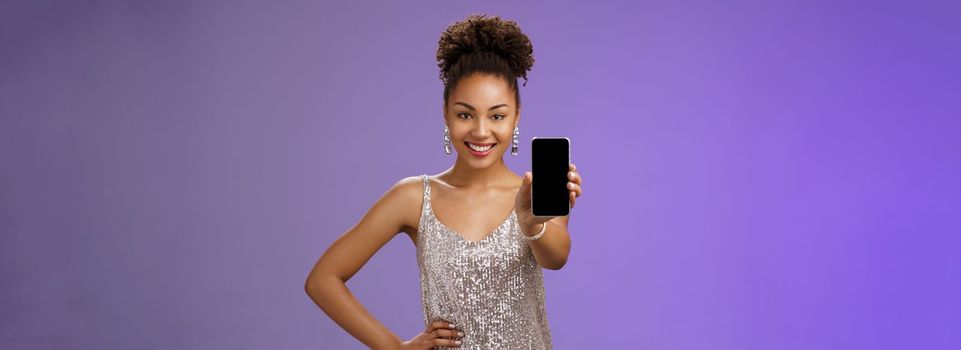 Sassy elegant african-american woman. in glittering shiny silver dress hold hand waist confident pose gladly smiling extend arm showing smartphone display check out cool app blue background.