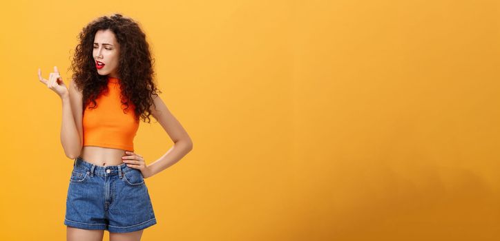 Portrait of moody arrogant and displeased european female with curly hairstyle and cropped top showing small and tiny object with fingers looking at little thing with disdain over orange background. Copy space