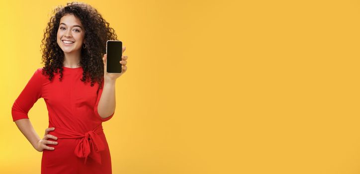 Girl brag with new phone she got on christmas feeling delighted holding mobile device in hand showing smartphone screena t camera, smiling broadly with uplifted mood over yellow background. Technology concept