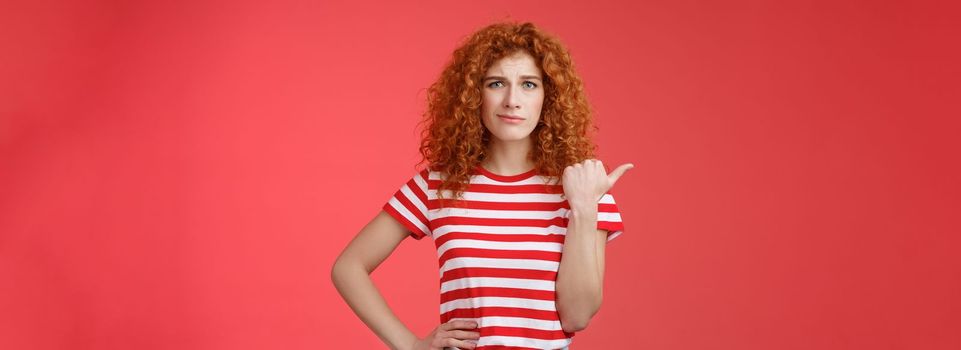 Girl not sure best choice. Doubtful redhead curly woman look suspicious frowning uncertain stare hesitant pointing left thumb unsure buy or not standing red background displeased. Copy space