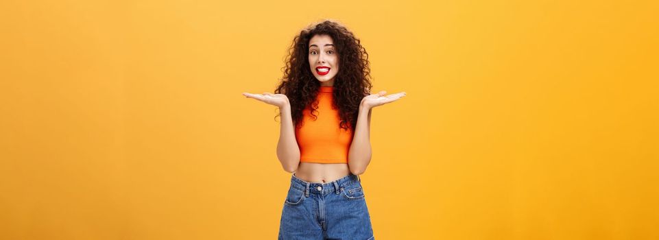 Oops honey sorry. Clueless silly stylish european female with curly hairstyle in cropped top shrugging with hands spread aside in unaware gesture smiling guilty and questioned over orange background.