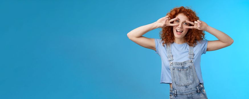 Lifestyle. Cheerful cute redhead ginger girl curly haircut show positivity peace victory signs eyes smiling broadly have fun playful silly childish mood wear denim summer overalls blue background.
