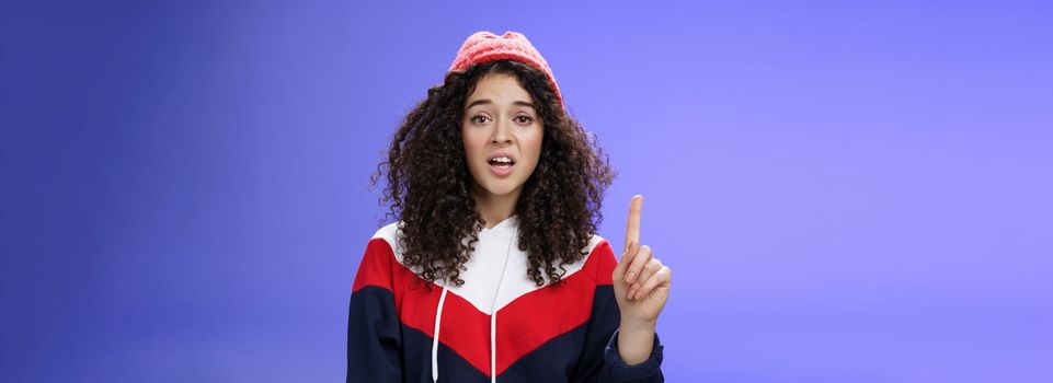 Rule number one do not mess with me babe. Portrait of swag and cool stylish young curly-haired woman in winter beanie showing index finger in prohibition or warning gesture over blue background.